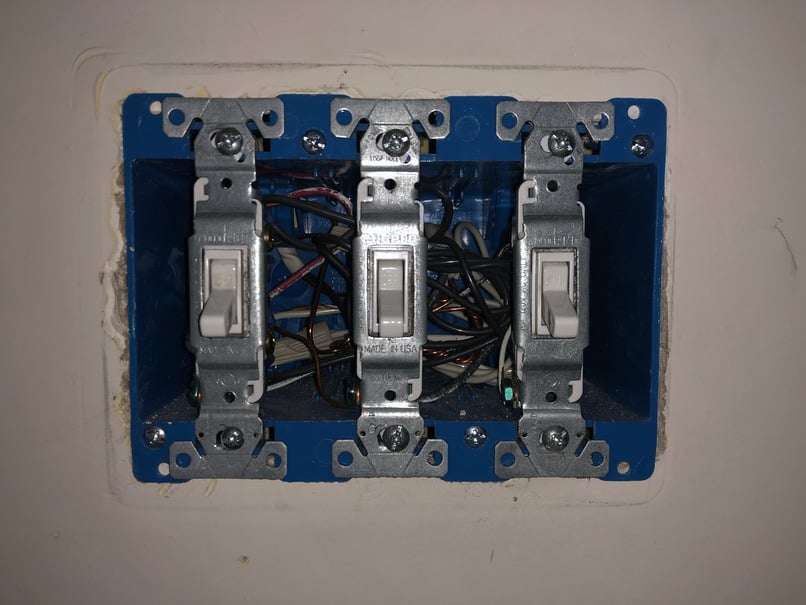 The unscrewed panel. Silver switches screwed to a blue container inset into the wall. Behind the switches is a jumble of wires.